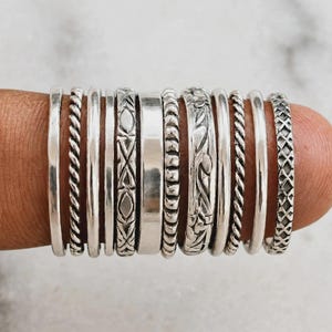 Puede incluir: Una colección de once anillos de plata dispuestos en fila, con diversas texturas y diseños que incluyen bandas lisas, patrones de cuerda trenzada, formas geométricas grabadas, una banda plana pulida y un motivo floral o de enredadera detallado.