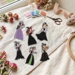 Puede incluir: Seis pegatinas troqueladas con figuras ilustradas con vestidos largos y sombreros de bruja. Los personajes visten trajes negros, morados y verdes, sosteniendo pequeñas calabazas o dulces. Dispuestos sobre una superficie de tela texturizada.