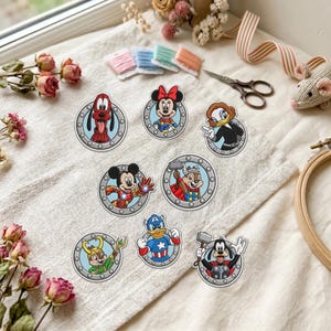 May include: A collection of eight circular embroidered patches featuring cartoon characters dressed as superheroes, including Mickey Mouse, Minnie Mouse, Goofy, Donald Duck, and Chip 'n Dale, each framed by a silver-colored metallic porthole design.