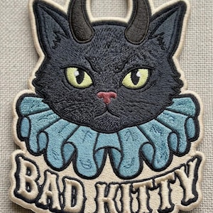 Puede incluir: Un parche bordado que muestra un gato negro con dos cuernos oscuros, ojos amarillos y nariz rosa. El gato lleva un cuello con volantes de color azul claro. Debajo del gato, el texto BAD KITTY está bordado en letras negras sobre fondo crema.