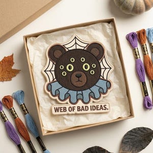 May include: An embroidered patch featuring a brown bear with six light green eyes, wearing a blue ruffled collar, set against a spider web background. Below the bear, a rectangular banner displays the text WEB OF BAD IDEAS in black letters.