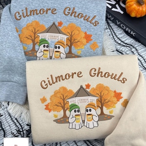 Op de afbeelding: Twee sweatshirts, één in heidegrijs en één in crème, elk voorzien van een borduursel van twee geesten met mutsen die koffiebekers vasthouden voor een prieel en herfstbomen, met de tekst Gilmore Ghouls in een boog erboven.