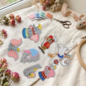 Puede incluir: Una colección de parches bordados que representan al elefante Dumbo y al ratón Timothy en varias posturas, incluyendo a Dumbo con grandes orejas rosas, una cigüeña cargando un bulto y diseños de circo con detalles en azul, rojo y amarillo.