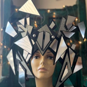 Op de afbeelding: Een zilveren en zwart geometrisch hoofddeksel met gespiegelde oppervlakken. Het hoofddeksel is gemaakt van meerdere driehoekige vormen en wordt gedragen op een mannequin.