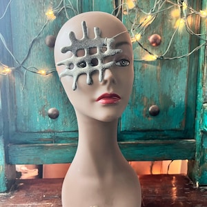 May include: A mannequin head with a silver-colored, abstract eye mask. The mask has a textured, web-like design and covers one eye. The mannequin has red lipstick and the word "Prima" is printed on the base.