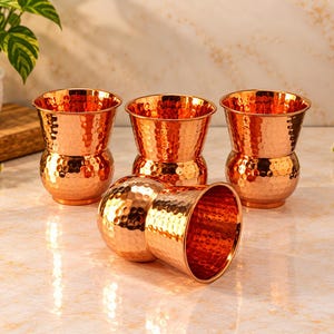 May include: A set of four copper cups with a distinct hammered texture. Each cup features a rounded base that tapers upward to a wider, circular rim. The polished metal surface reflects light, highlighting the indented pattern across the exterior.