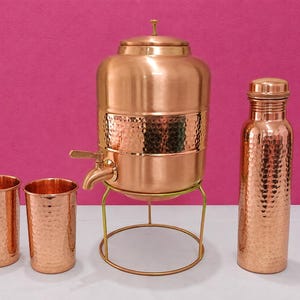 May include: A copper beverage set consisting of a large hammered water dispenser with a spigot on a metal stand, two matching hammered tumblers, and a tall hammered water bottle with a screw-top lid, all featuring a polished metallic finish.