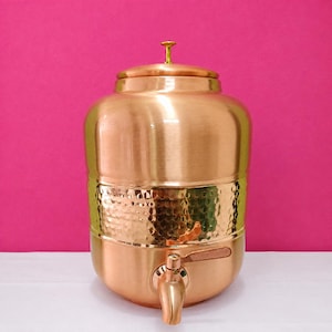 May include: A large, cylindrical copper water dispenser featuring a smooth upper section and a wide, hammered texture band around the middle. It includes a matching copper lid with a small gold-toned knob on top and a functional copper spigot at the base.