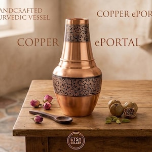 Op de afbeelding: Een koperen vat met een taps toelopende hals en een brede basis, voorzien van donkere, ingewikkelde bloemengravures op de hals en het lichaam. De tekst HANDCRAFTED AYURVEDIC VESSEL en COPPER ePORTAL is zichtbaar. Een houten lepel, gedroogde rozenknoppen en zaden liggen in de buurt.