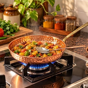 May include: A round, hammered copper frying pan with a long, polished brass handle sits on a gas stove burner. Inside the pan, a mixture of sautéed potatoes, tomatoes, and green peppers is being cooked, with a silver serving spoon resting on top.