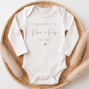 May include: A white long-sleeve baby bodysuit featuring the text PROMOTED TO Nan + Pop IN 2027 in a tan font, with a small tan heart icon centred below the text. The garment has a round neckline and three snaps at the bottom for closure.