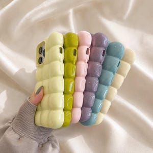 May include: A collection of five smartphone cases featuring a raised, puffy, quilted texture. The cases are available in pastel yellow, lime green, soft pink, lavender, and dusty blue, each with a glossy finish and a cutout for a camera lens.