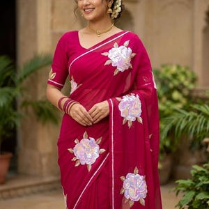 May include: A vibrant magenta saree featuring large, white, sequined floral appliqués with gold-toned leaf embroidery. The garment includes a matching short-sleeved blouse with similar floral detailing on the sleeves and a thin white border.