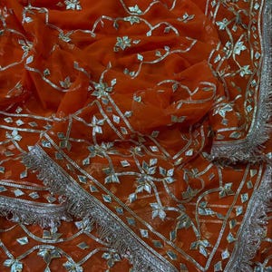 May include: A vibrant orange fabric featuring intricate silver metallic embroidery and reflective mirror-like accents arranged in floral patterns. The edges are finished with a decorative silver fringe border and detailed geometric silver trim.