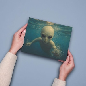 May include: A rectangular canvas print featuring a high-resolution image of a pale, large-headed extraterrestrial figure swimming underwater. The alien has dark, almond-shaped eyes and is surrounded by deep blue water with visible light refraction.