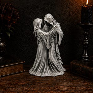 K&ouml;nnte beinhalten: Eine silberfarbene Skulptur, die zwei kapuzentragende Gestalten in langen, flie&szlig;enden Gew&auml;ndern zeigt, die sich umarmen und tanzen. Die Figuren haben skelettartige H&auml;nde und sind in Stoff mit tiefen Falten geh&uuml;llt, in einer tanzenden Pose.