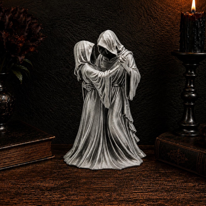 K&ouml;nnte beinhalten: Eine silberfarbene Skulptur, die zwei kapuzentragende Gestalten in langen, flie&szlig;enden Gew&auml;ndern zeigt, die sich umarmen und tanzen. Die Figuren haben skelettartige H&auml;nde und sind in Stoff mit tiefen Falten geh&uuml;llt, in einer tanzenden Pose.