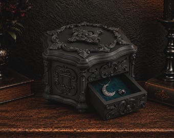 Könnte beinhalten: Eine mattschwarze Schmuckschatulle mit verzierten, erhabenen Schnörkeln auf dem Deckel und den Seiten. Die Schatulle verfügt über eine ausziehbare Schublade mit dunkelblaugrünem Samtfutter, in der ein silberner Mondanhänger und ein Ring liegen.