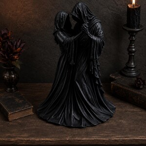 K&ouml;nnte beinhalten: Eine schwarze Skulptur, die zwei kapuzenbedeckte Gestalten in langen, flie&szlig;enden Gew&auml;ndern zeigt, die eng beieinander stehen. Die Figuren sind in dunklen, strukturierten Stoff mit tiefen Falten geh&uuml;llt, ihre Gesichter sind von Kapuzen verdeckt.
