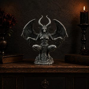 May include: A silver-toned statue of a winged, horned figure with a goat-like head and human-like torso, depicted in a seated, crouching position with arms extended outwards. The figure features detailed wings, textured skin, and a circular emblem on the chest.
