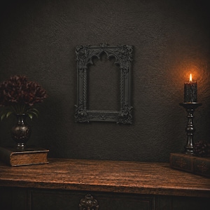 May include: A rectangular black picture frame with ornate gothic-style carvings, featuring a pointed arch top and twisted column side details. The frame is mounted on a dark textured wall, flanked by a dark vase with flowers and a lit black candle.
