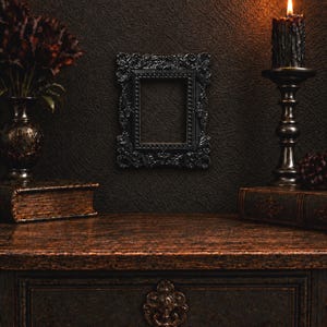 May include: A rectangular black picture frame with an ornate, embossed border featuring intricate scrollwork and bead detailing. The frame is mounted on a dark, textured wall between a vase of dark flowers and a lit black candle in a metal holder.