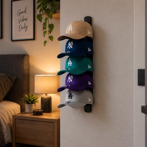 May include: A vertical black wall-mounted rack holding five baseball caps in tan, navy blue, teal, purple, and light grey. Each cap features a white embroidered logo on the front, including the LA and NY insignias, arranged in a single column.