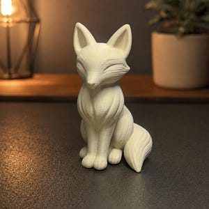 Op de afbeelding: Een klein, gebroken wit, 3D-geprint vossenbeeldje in zittende houding. De vos heeft puntige oren, gesloten ogen en een textuur met zichtbare lagen. De staart is om het lichaam gekruld en het beeldje heeft een matte, monochrome uitstraling.