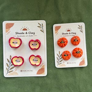 Könnte beinhalten: Zwei Präsentationskarten von Shade & Clay Handmade art studio. Die linke Karte zeigt vier rote apfelförmige Knöpfe mit gelber Mitte. Die rechte Karte zeigt vier orangefarbene kürbisförmige Knöpfe. Jeder Knopf hat zwei Löcher zum Annähen.