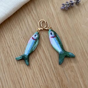 Op de afbeelding: Een paar hangende oorbellen met handbeschilderde visbedels in groen, blauw en roze. Elke vis heeft een klein oog en een rode mond, hangend aan goudkleurige metalen klapoorhaken die de oorbellen op hun plek houden tijdens het dragen.