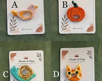 Può includere: Quattro portatovaglioli in argilla fatti a mano su cartoncini espositivi etichettati A, B, C e D. L'opzione A è un uccellino rosa, B una zucca arancione, C una lumaca color ottanio e oro, e D un muso di gatto tigrato arancione con un ciondolo a pesce.