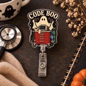 May include: A badge reel featuring a ghost character standing behind a red medical crash cart with a heart monitor, a stethoscope, and an oxygen tank. The text CODE BOO is printed in black at the top. The design is on a clear acrylic base with a clip.