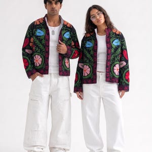 Pode incluir: Duas jaquetas pretas com zíper apresentando bordados florais multicoloridos vibrantes em tons de rosa, azul, laranja e vermelho. Cada jaqueta é usada sobre uma regata branca simples, combinada com calças brancas largas e calçados bege sem cadarço.