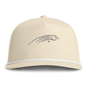 May include: A cream-colored baseball cap featuring a black line art graphic of a shooting star with a trail and small four-pointed stars. The cap includes a white cord trim running horizontally across the base of the crown above the brim.
