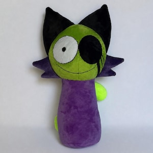 Puede incluir: Un peluche con cara verde, un ojo blanco y negro, un parche negro sobre el otro ojo y orejas negras puntiagudas. El cuerpo es morado con pequeños detalles en verde lima a los lados y una sonrisa cosida en la cara verde.