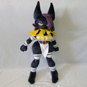 Puede incluir: Un muñeco de peluche que representa un personaje felino negro con grandes orejas puntiagudas, ojos amarillos y cabello morado. La figura lleva un cuello amarillo y blanco, un cinturón azul con un emblema circular rojo y vendas blancas.