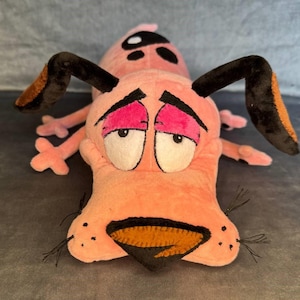 May include: A pink plush dog toy featuring large white eyes with black pupils, dark brown floppy ears, a dark brown nose, and black whiskers. The character has a worried expression with pink eyelids and black spots on its back and head.