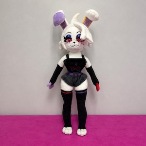 Könnte beinhalten: Eine Plüsch-Hasenpuppe mit weißem Fell, roten Augen und langen Ohren mit violetten und gelben Akzenten. Die Puppe trägt ein schwarzes Tanktop mit einem roten Stern-Motiv, eine schwarze Shorts mit violettem Gürtel sowie schwarze Armstulpen und Strümpfe.