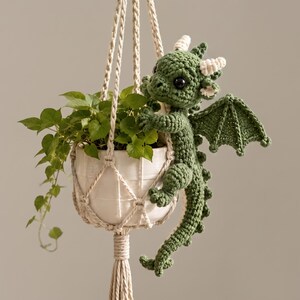 Puede incluir: Un dragón de peluche tejido a ganchillo en color verde con cuernos y alas blancas, aferrado a una maceta de cerámica blanca sostenida por un colgador de macramé color crema. Una pequeña planta verde crece dentro de la maceta con hojas colgantes.
