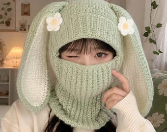 May include: A light green crochet balaclava featuring long, floppy rabbit ears and two white flowers with yellow centers attached to the sides. The knit headgear covers the head and neck, leaving an opening for the eyes and nose area.
