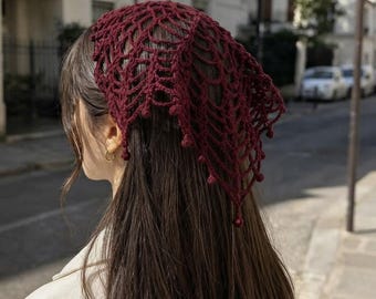 May include: A deep burgundy triangular headscarf crafted from yarn in an open, web-like crochet pattern. The accessory features a delicate, intricate network of interconnected loops and knots that form a geometric design, ending in small, rounded decorative beads at the points along the bottom edge.
