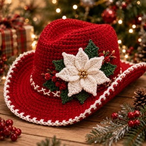 Op de afbeelding: Een rode gehaakte cowboyhoed met een witte gehaakte kerststerbloem in het midden, geaccentueerd met groene gehaakte bladeren en kleine rode bessen. De rand en de kroon zijn afgewerkt met een witte gestikte rand.