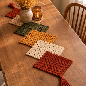 May include: A table runner composed of six connected square crochet motifs in shades of rust, cream, and olive green. The runner features a repeating openwork pattern and is finished with a decorative tassel at each end, arranged in a linear sequence.