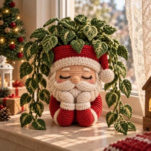 Könnte beinhalten: Ein gehäkelter Weihnachtsmann-Übertopf mit roter Mütze und Mantel sowie weißem Bart und Besatz. Der Übertopf enthält eine hängende Grünpflanze mit panaschierten Blättern. Die Figur hat geschlossene Augen und die Hände unter dem Kinn.