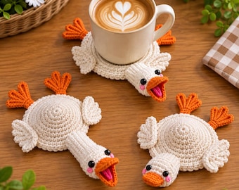 May include: Three handmade crochet coasters shaped like white ducks with orange webbed feet and beaks. Each coaster features a round body, two wings, and a duck head with black eyes and pink cheeks. One coaster supports a ceramic mug of coffee.