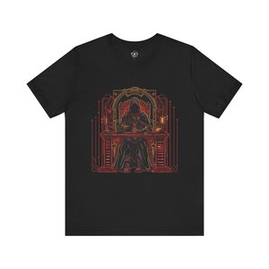 May include: A black short-sleeve t-shirt featuring a graphic print of a hooded figure sitting at a desk. The design uses red and orange lines to depict a throne-like chair, electronic equipment, and a circular emblem on the desk in a dark setting.