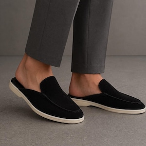May include: A pair of black suede slip-on mules featuring a moccasin-style stitched toe seam and a flat, off-white rubber sole. The footwear is paired with grey trousers, showcasing a backless design that leaves the heel exposed for easy wear.