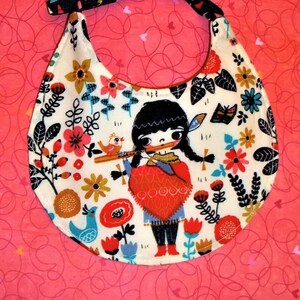May include: A baby bib with a white background featuring a colorful floral pattern and a cartoon illustration of a girl playing a flute. The bib has a black and patterned neck strap and is set against a pink background.