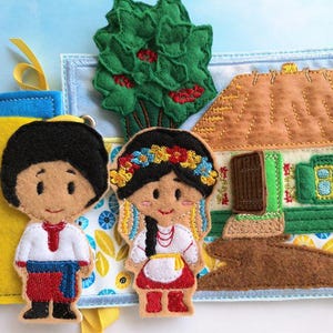May include: A felt quiet book page featuring a Ukrainian theme. It includes felt figures of a boy and girl in traditional clothing, a house with a thatched roof, and a green tree. The background is light blue with floral patterns.