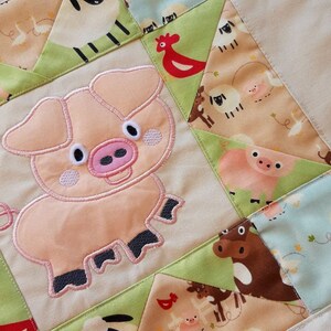 May include: A beige quilted fabric square with a pink border. The fabric is decorated with a patchwork design featuring various farm animals, including pigs, sheep, cows, and chickens. The animals are embroidered in pink, black, and white.